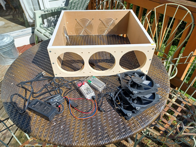 A wicker table on a deck holds a wooden frame with three round holes on one side, three computer fans, some electronics, three grills, and some machine screws and nuts.