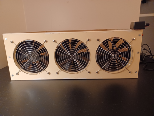 Three steel grills, consisting mostly of concentric circles, cover the large holes in a wooden frame. Fans show through the holes.