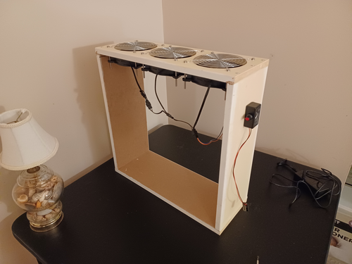 A wooden frame with electronics attached at the top and sides sits near a small lamp atop a black card table.