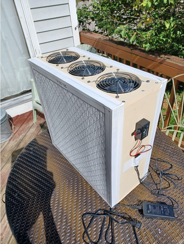 A Corsi-Rosenthal box the size of a tower computer sits atop a wicker table on a deck, along with its power supply. The wood is painted cream color. The filters and tape are white. The grills are steel, and the electronics are mostly black.